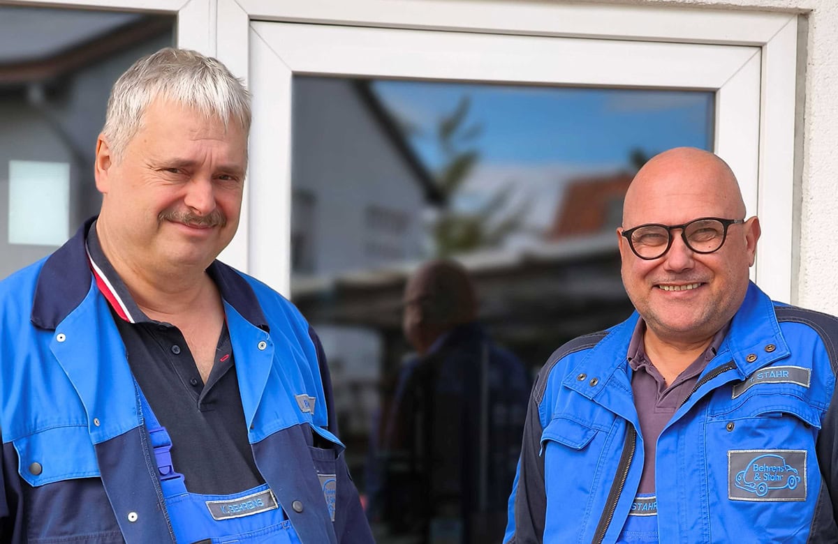 Zwei Männer in blauen Arbeitsjacken lächeln vor einem Fenster.